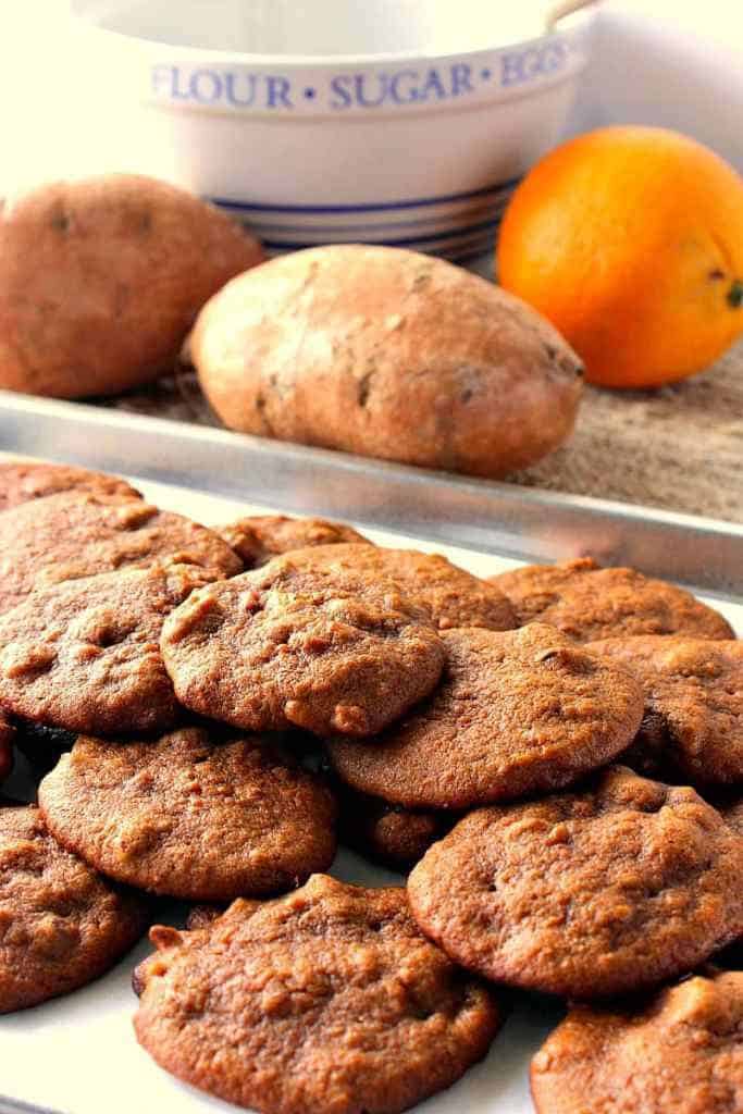 Tray of Amish sweet potato cookies in the foreground and sweet potatoes and an orange in the background.