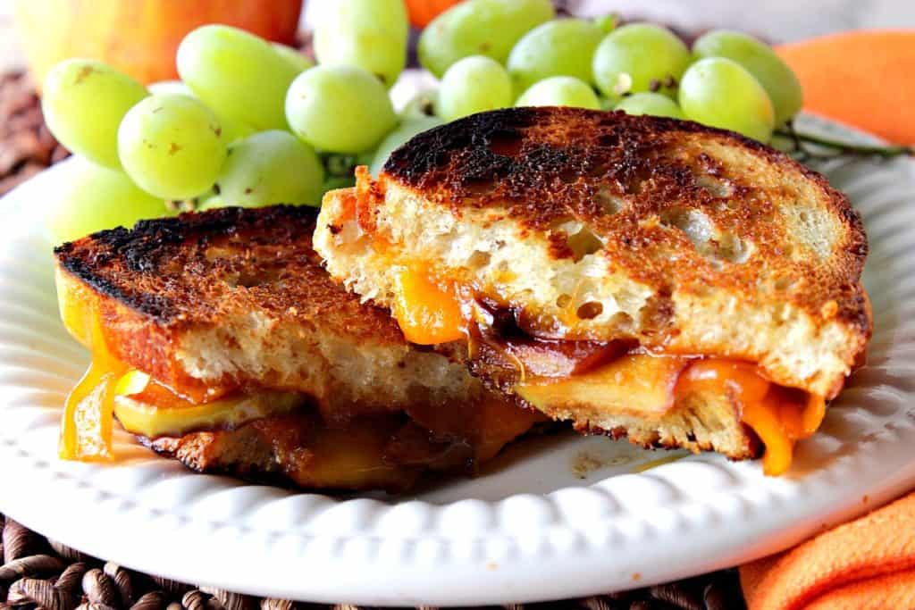 A horizontal photo of a grilled cheese sandwich with caramelized apples on a white plate with an orange napkin and green grapes.