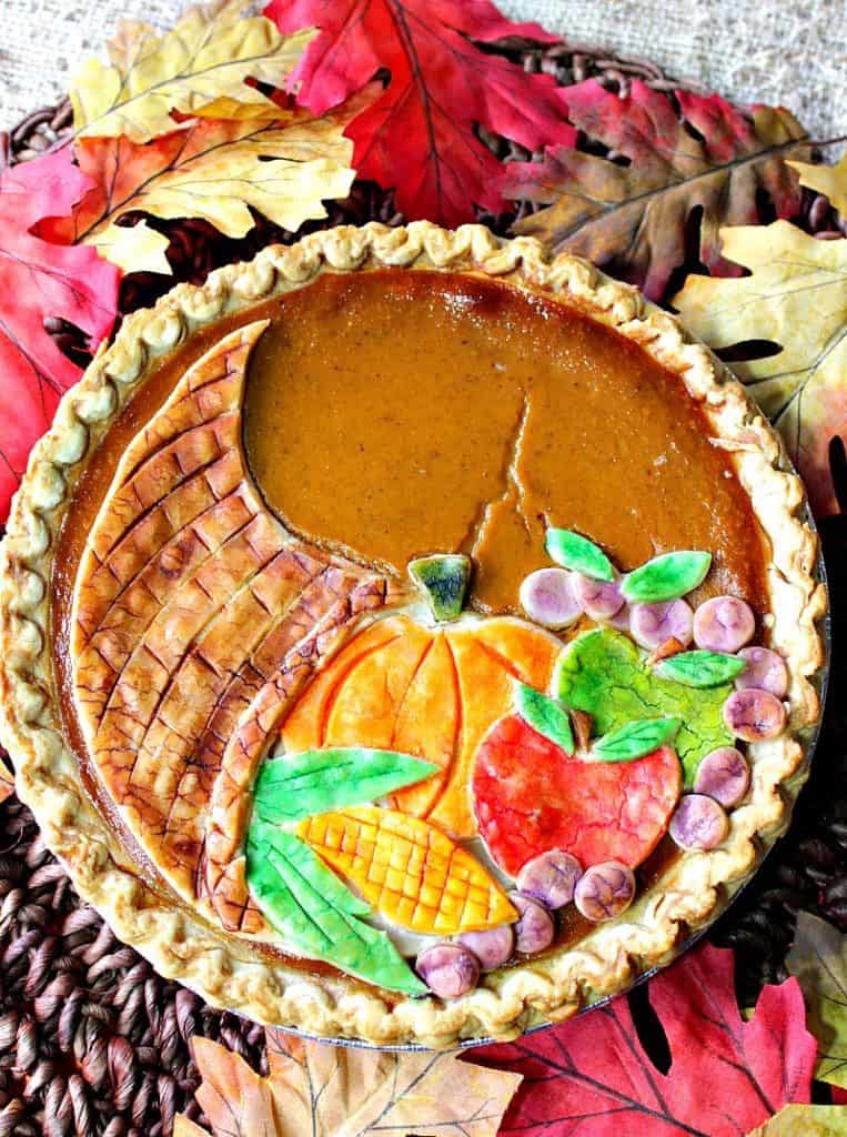 Overhead closeup photo of a colorful painted cornucopia pumpkin pie with corn, apples, grapes, and a pumpkin.