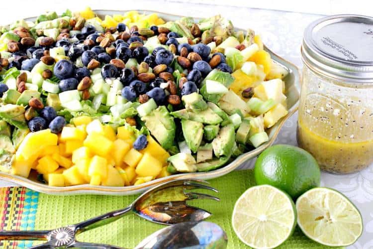 Vertical photo of a heart healthy avocado and mango salad on a platter with lime vinaigrette and blueberries.