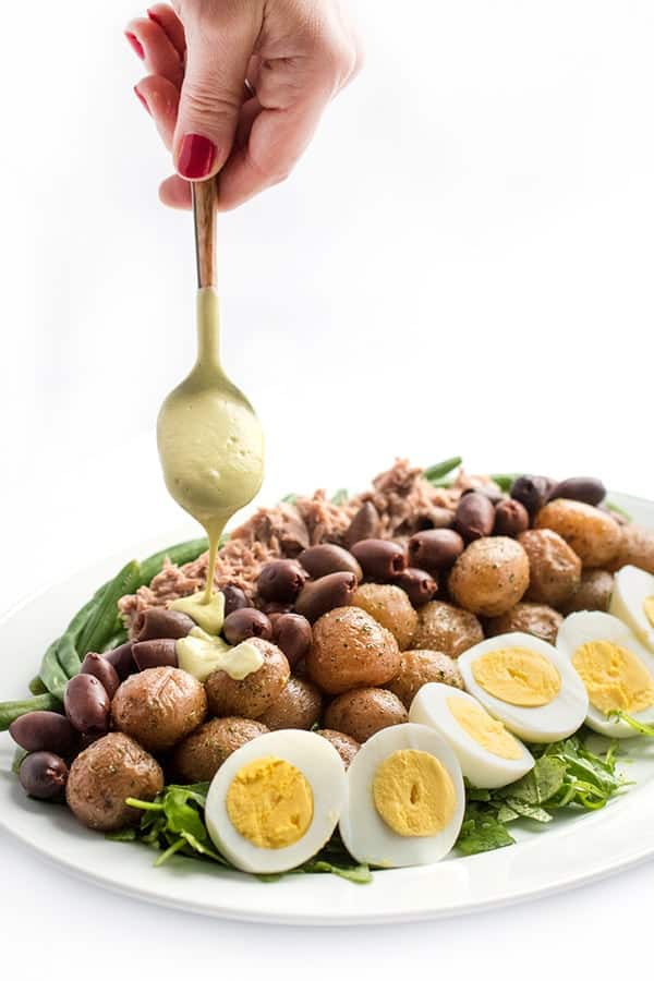 A hand holding a salad dressing spoon drizzling dressing over potato and egg salad with olives for healthy salad recipe roundup.