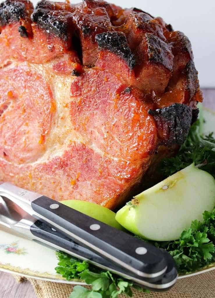 A closeup vertical photo of a mustard apricot glazed ham with apples and carving utensils on a plate. Christmas dinner recipe roundup.
