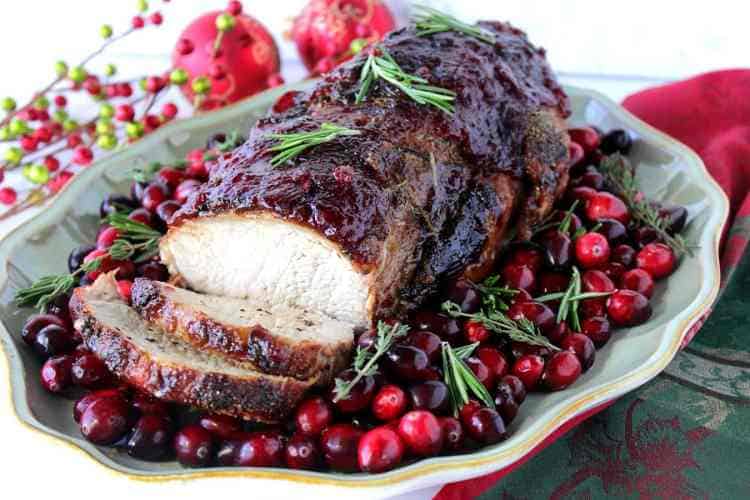 A horizontal photo of a sliced cranberry glazed pork roast on a platter with whole cranberries and fresh herbs. Christmas dinner recipe roundup.
