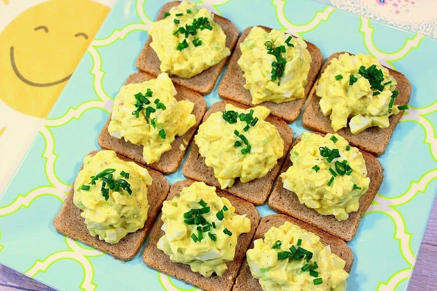 A horizontal photo of egg salad canapes on a blue and yellow plate with a sunshine napkin on the side.