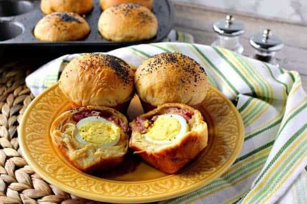 Hard boiled eggs wrapped in a biscuit with ham and cheese on a golden color plate with a muffin tin in the background.