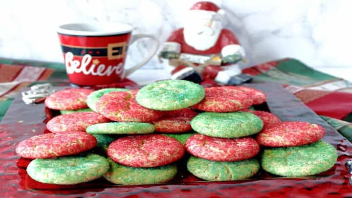 A red glass plate filled with Santa's Favorite Sugar Cookies in red and green along with a coffee mug in the background.