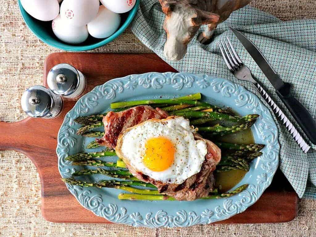 Overhead picture of steak, egg, and asparagus on a blue oval plate with a bowl of eggs in the background.