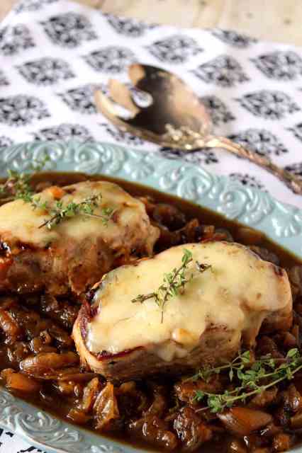 A vertical closeup photo of French onion pork chops on a blue platter with caramelized onions, melted cheese and fresh thyme as garnish.