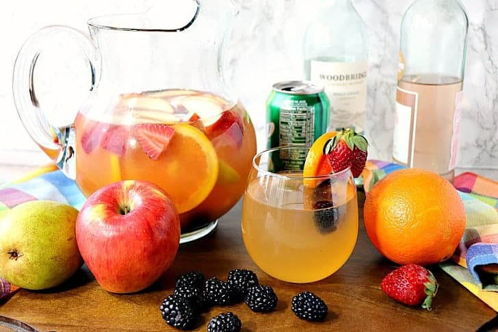 A glass of sangria with blackberries, apples, and oranges with a pitcher in the background along with a few bottles of wine.