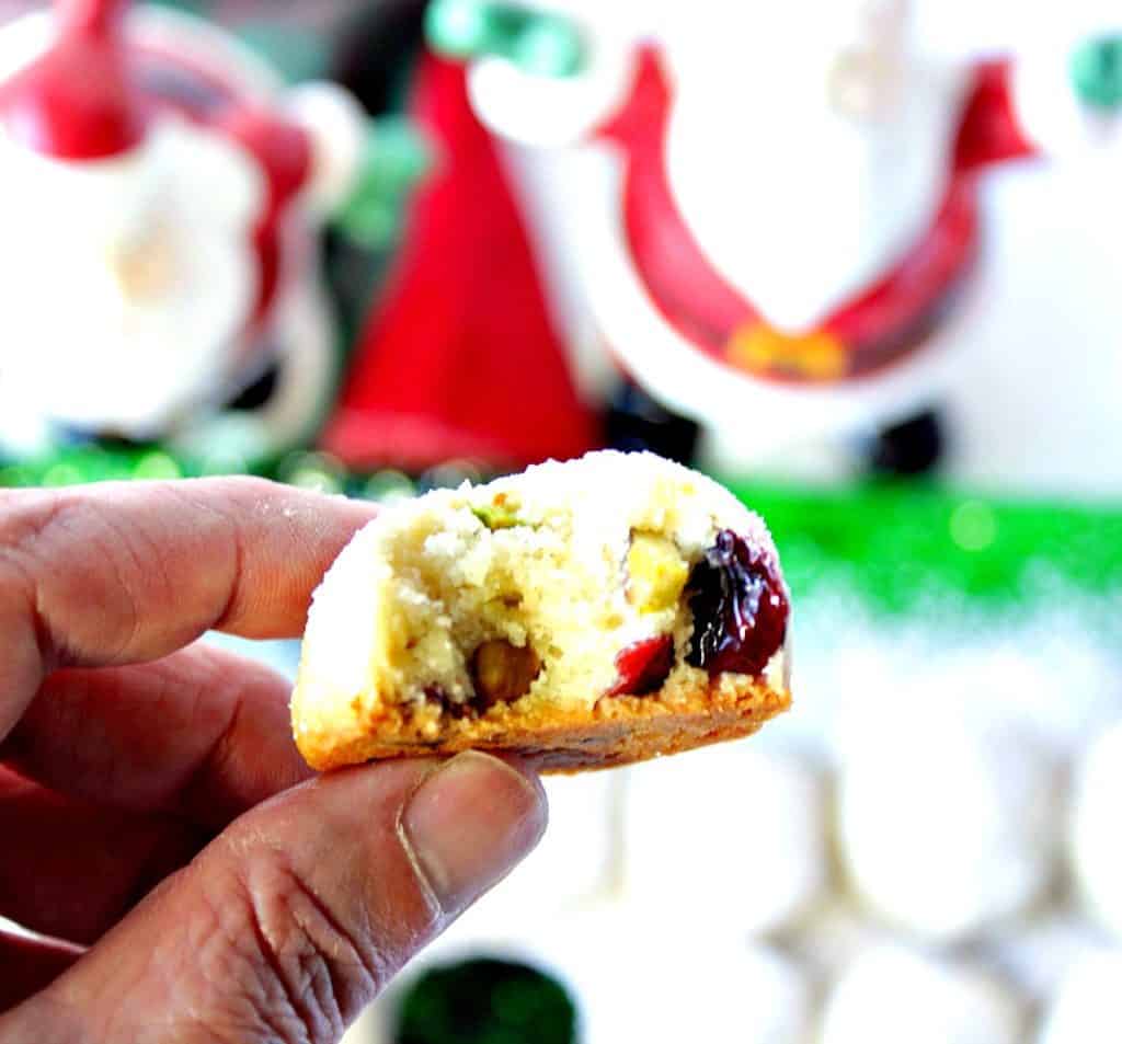 A hand holding a snowball cookie with a bite taken out and you can see the pistachios and cranberries.