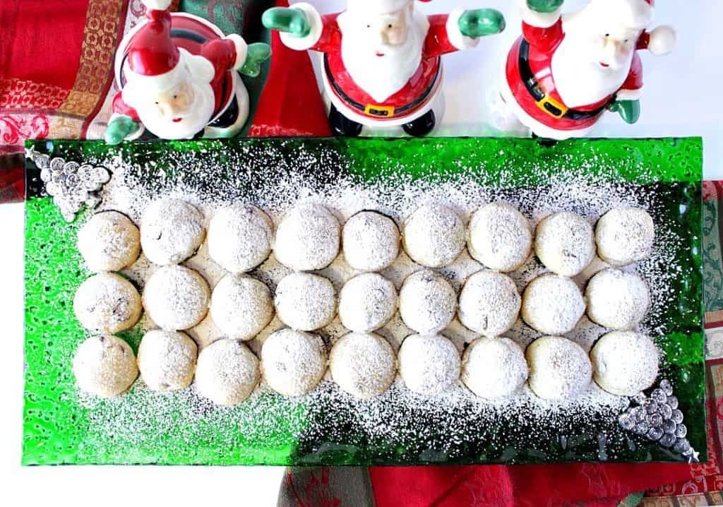 An overhead photo of a green glass Christmas plate filled with Snowball Cookies with Pistachios and Cranberries with a confectioners sugar dusting.