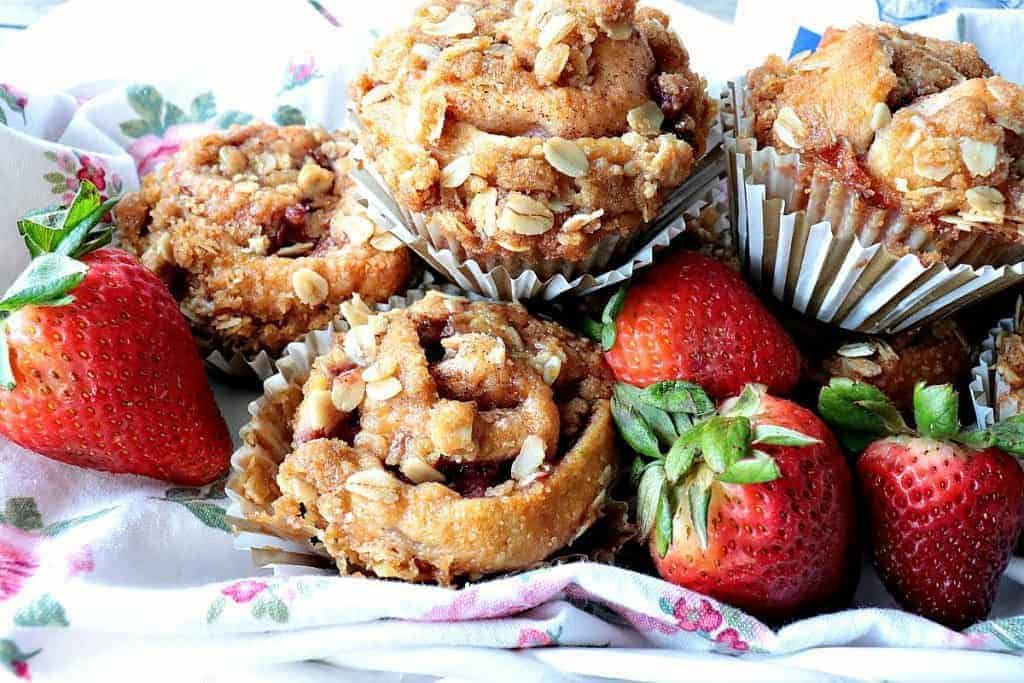 Strawberry muffins piled up on a pretty floral pink napkin with fresh strawberries surrounding the muffins.