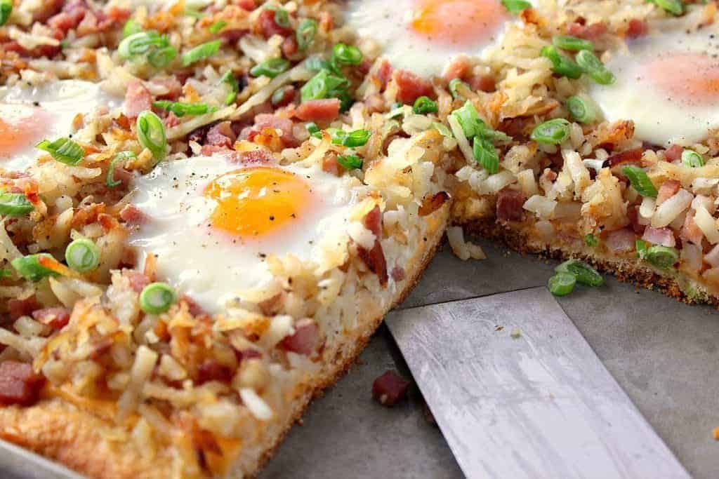 Closeup photo of a hash brown breakfast pizza with a slice taken out and over easy eggs on top.