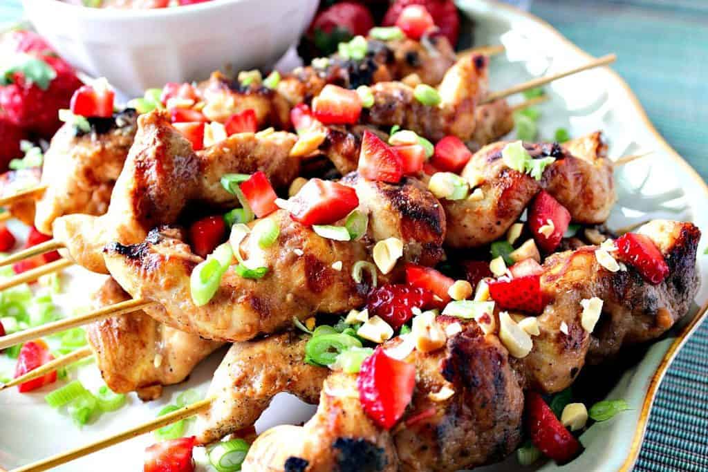 Horizontal closeup photo of chicken skewers on a plate with red strawberries and green scallions.