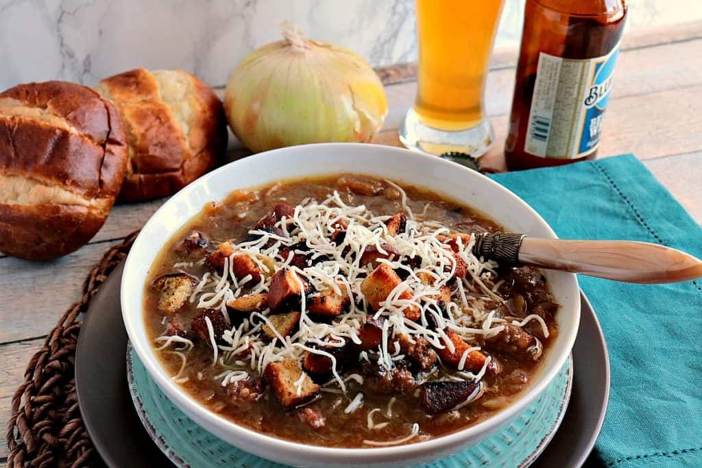 A bowl of German onion soup with onions and pretzel roll in the background along with a glass of beer for Soup, Stews, and Chowder Recipes.