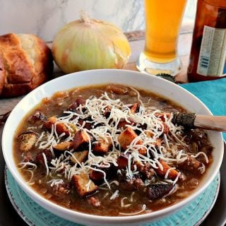 A bowl of German onion soup with onions and pretzel roll in the background along with a glass of beer.