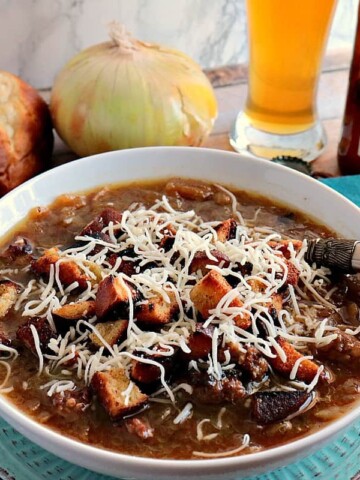 A bowl of German onion soup with onions and pretzel roll in the background along with a glass of beer.