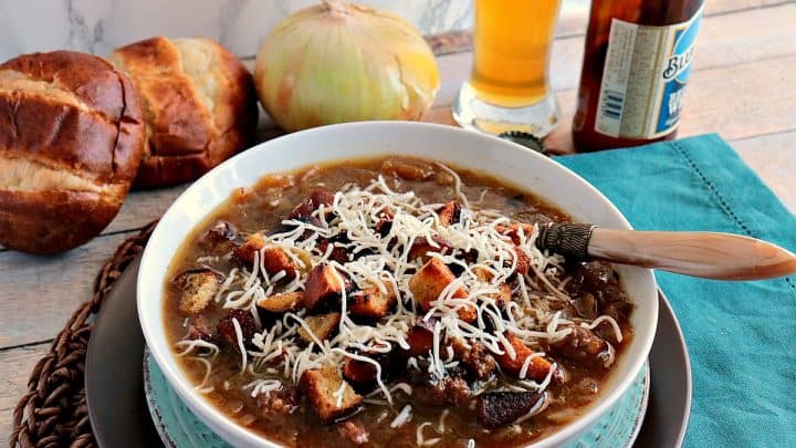 A bowl of German onion soup with onions and pretzel roll in the background along with a glass of beer.