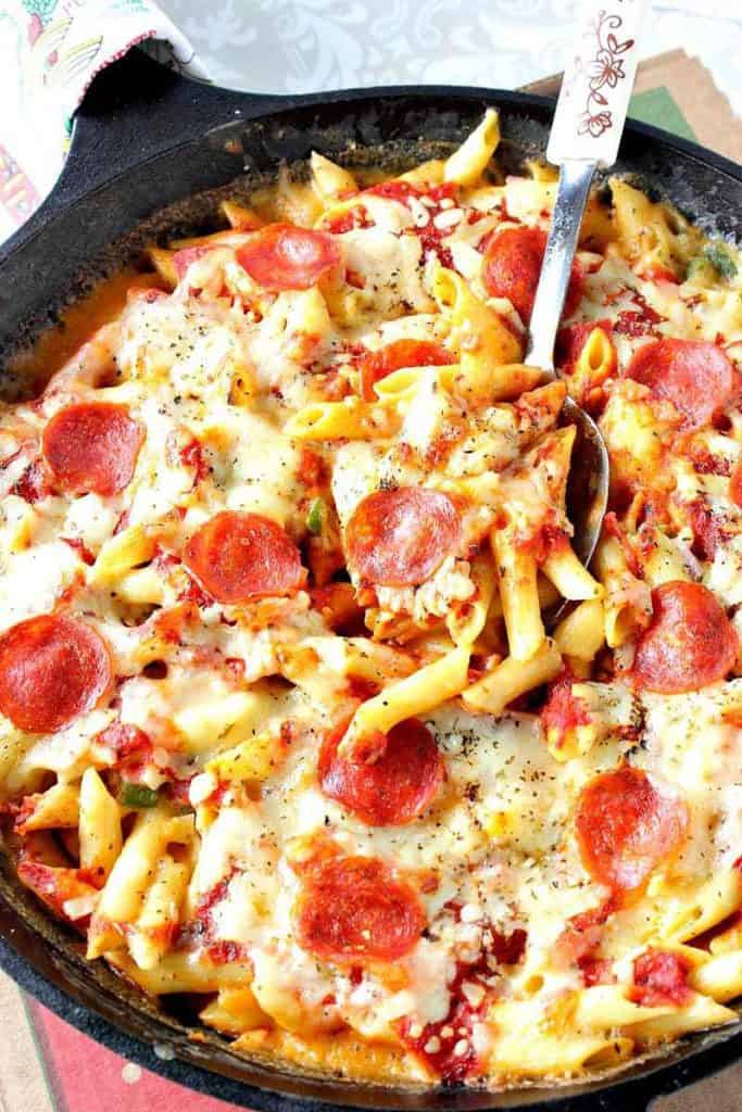 Vertical closeup image of pepperoni pizza mac and cheese in a skillet with a serving spoon and lots of melted cheese.