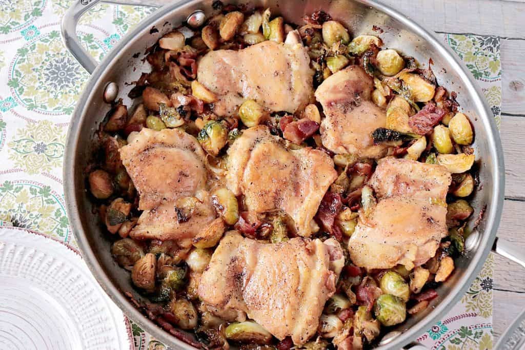 Chicken thighs in a skillet with bacon and Brussels sprouts.