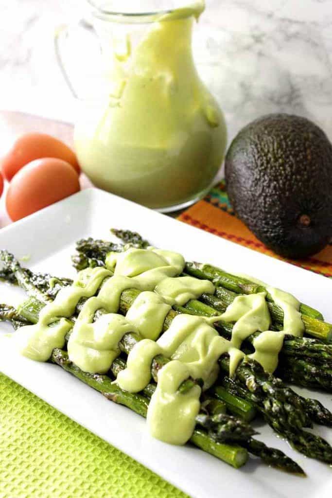 A vertical photo of asparagus on a rectangle white plate smothered in avocado hollandaise sauce.