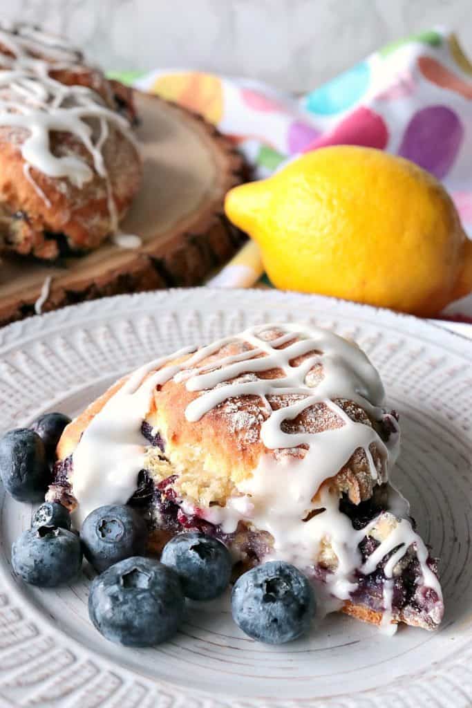 Vertical image of a fresh blueberry scone in the foreground and a muti-color polka dot napkin in the background with a lemon.