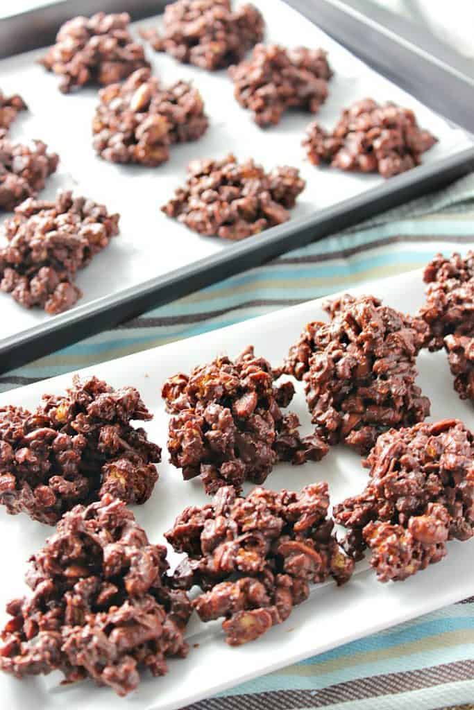 A vertical photo of a plate of peanut butter granola jumbles in the foreground and a tray of them in the background.