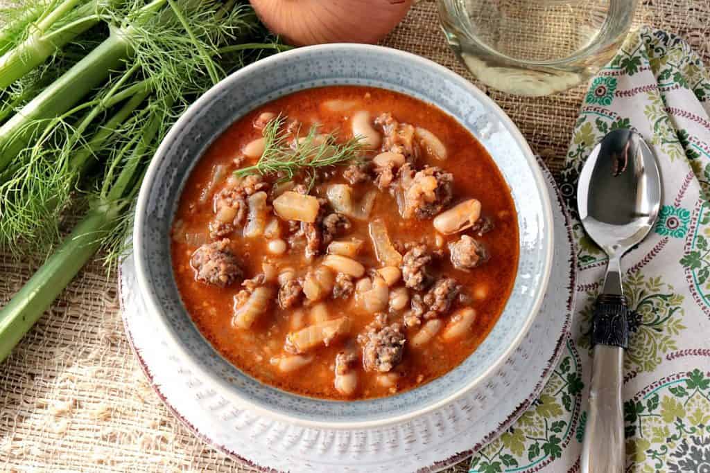 This hearty White Bean, Fennel & Italian Sausage Soup is sure to please the entire family. The depth of flavor from the blend of sweet fennel and the white beans is a sumptuous treat for the taste buds. - Kudos Kitchen by Renee A hearty bowl of White Bean, Fennel & Italian Sausage Soup with Fennel, Onion and a glass of wine.