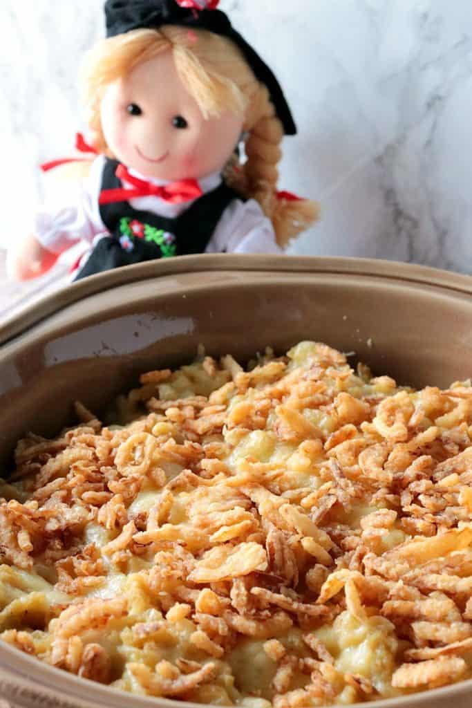 A closeup vertical image of German spaetzle casserole in a brown round dish with a little German doll in the background