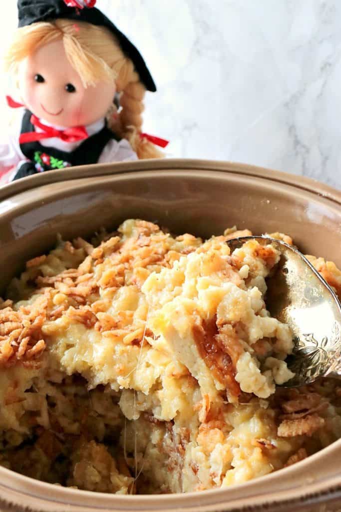 Closeup vertical photo of a casserole dish filled with layered spaetzle casserole with cheese and crispy onions with a German doll in the background.