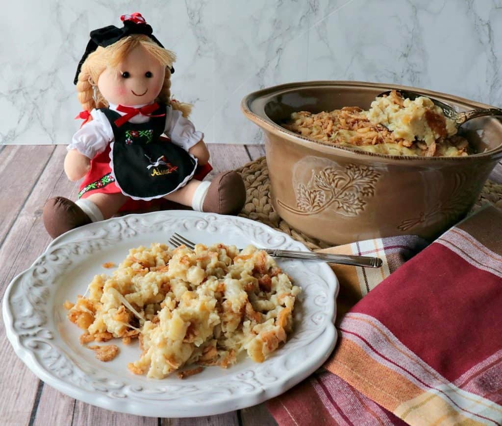 Buttered German egg dumplings (spaetzle) are layered in a casserole dish along with shredded Gruyere cheese and crispy, crunchy onions to bring you this delicious recipe for Layered German Spaetzle Casserole with Cheese & Crunchy Onions. - www.kudoskitchenbyrenee.com A plate filled with German spaetzle casserole with a little German doll in the background.