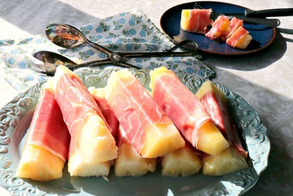 Many Pineapple spears on a blue blue plate wrapped in prosciutto ham.