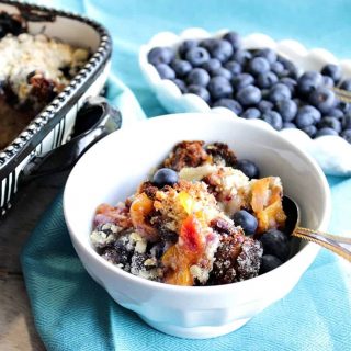 A horizontal photo of a dish of Blueberry Peach Crisp in the foreground along with a bowl of fresh blueberries in the background.