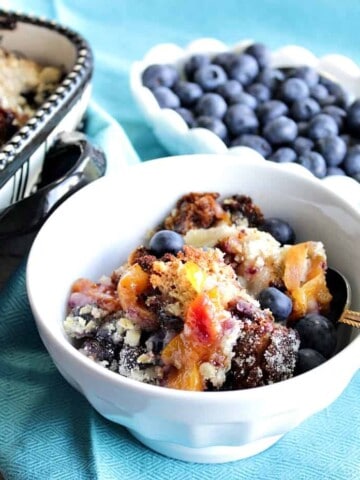 A horizontal photo of a dish of Blueberry Peach Crisp in the foreground along with a bowl of fresh blueberries in the background.