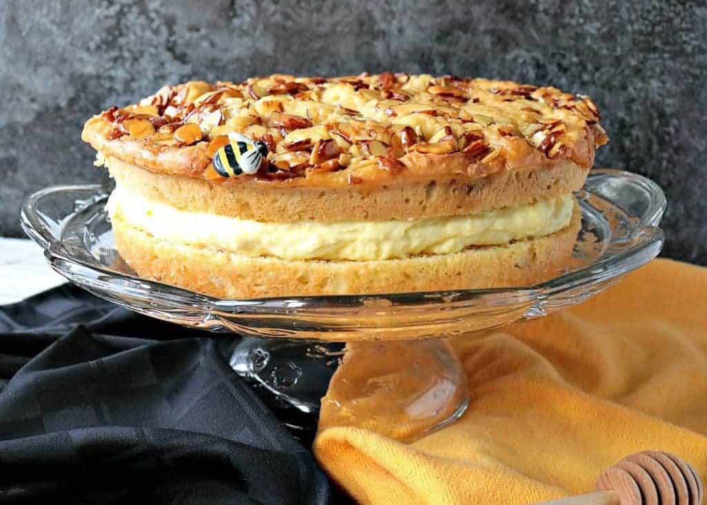 Layered German Bee Sting Cake on a glass cake plate with a black and yellow napkin under the stand.