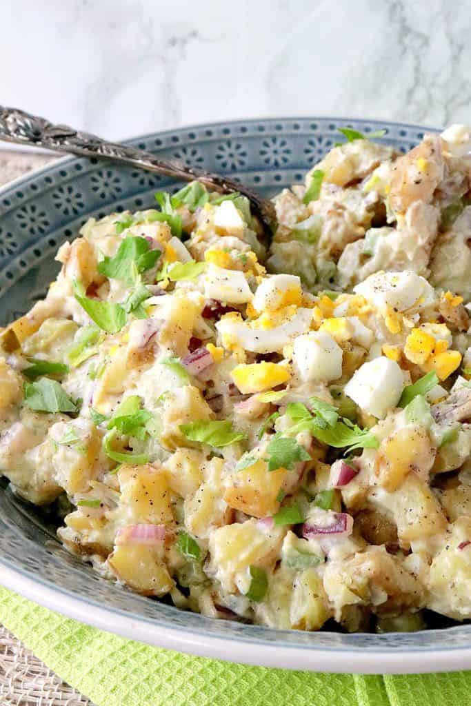 A closeup photo of a blue bowl of Dijon potato salad with a spoon, hard boiled eggs, and parsley.