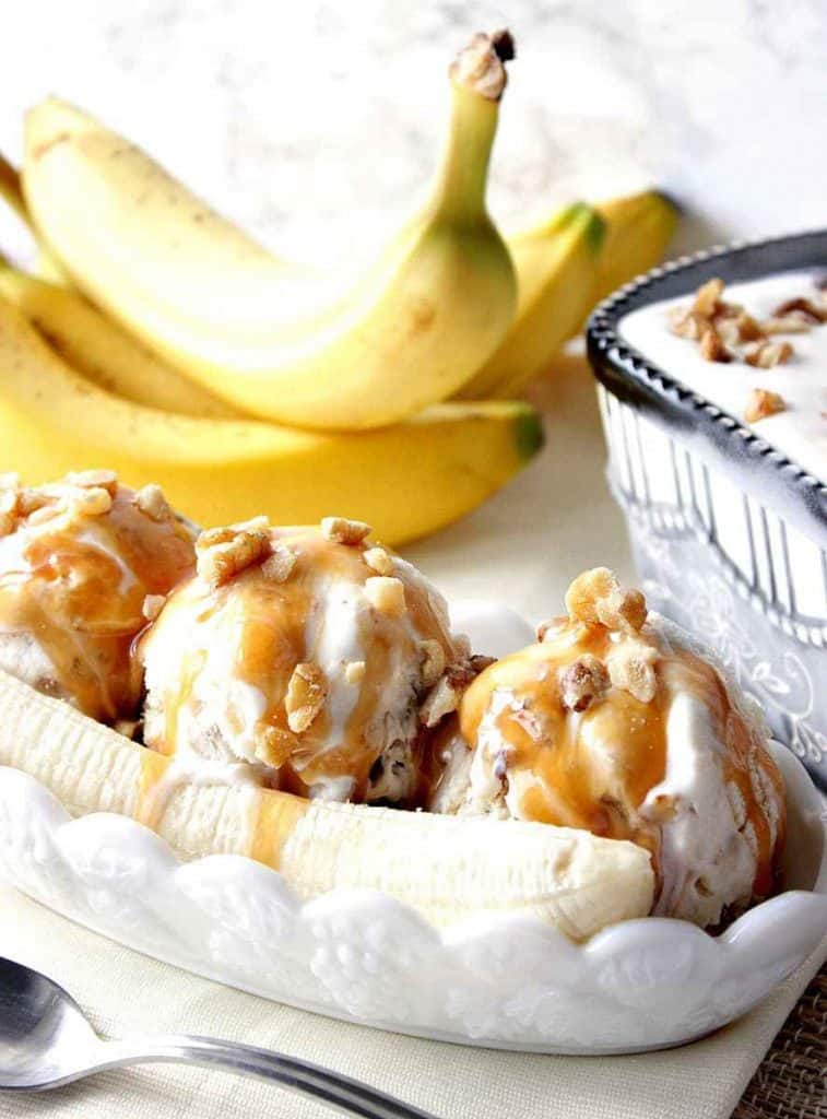 Vertical closeup photo of a dish of banana walnut ice cream with bananas in the background and carmel sauce on top.