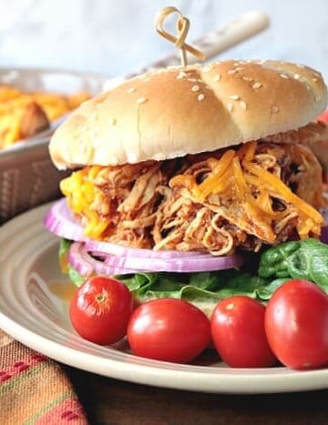 A shredded salsa chicken sandwich on a plate with tomatoes and a dish of salsa chicken in the background.
