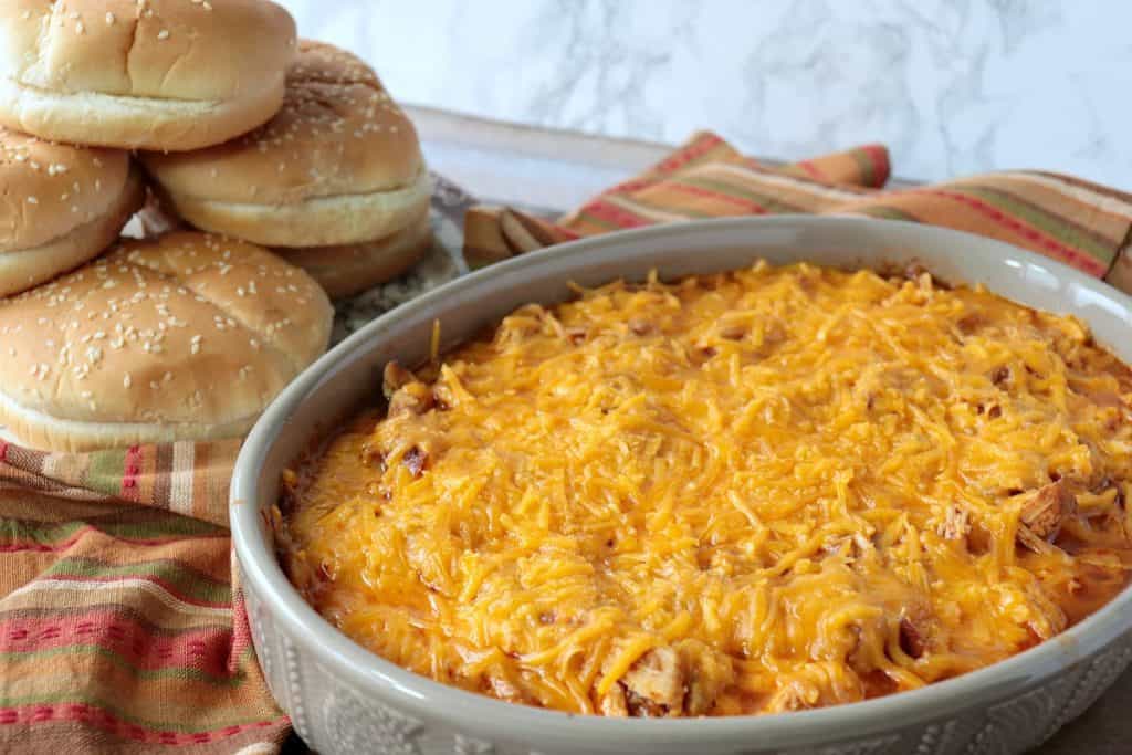 An casserole dish filled with shredded salsa chicken topped with tons of melted cheddar cheese and a pile of buns in the background.