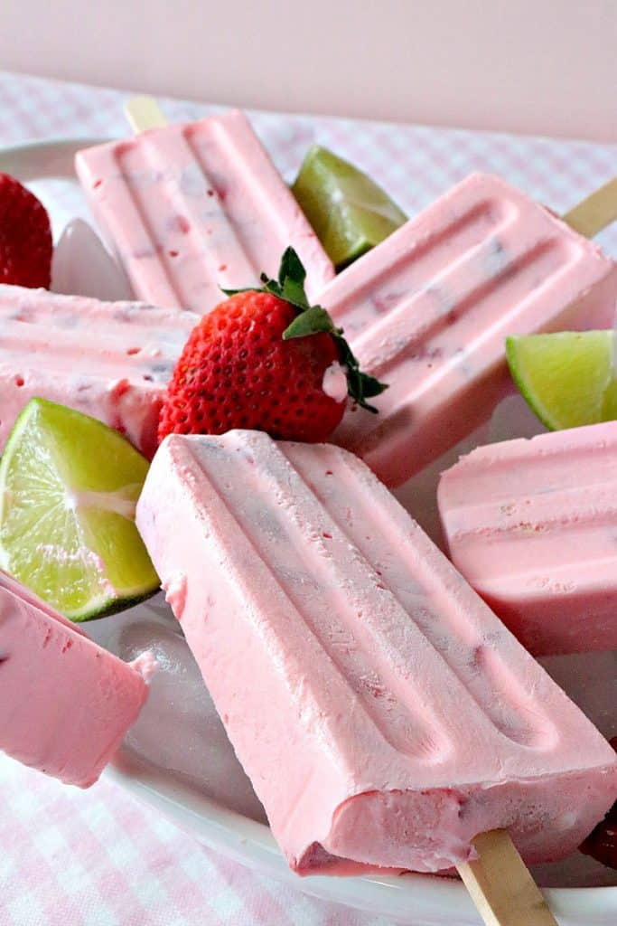 Closeup picture of a plate of whipped cream strawberry popsicles with lime wedges and strawberries on a pink checked tablecloth.