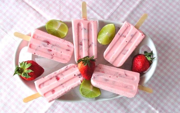 Overhead picture of a tray of strawberry cream popsicles with strawberries and lime wedges.