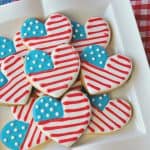 A horizontal photo of American Flag Heart decorated cookies on a square white plate with a red and white checked napkin underneath.
