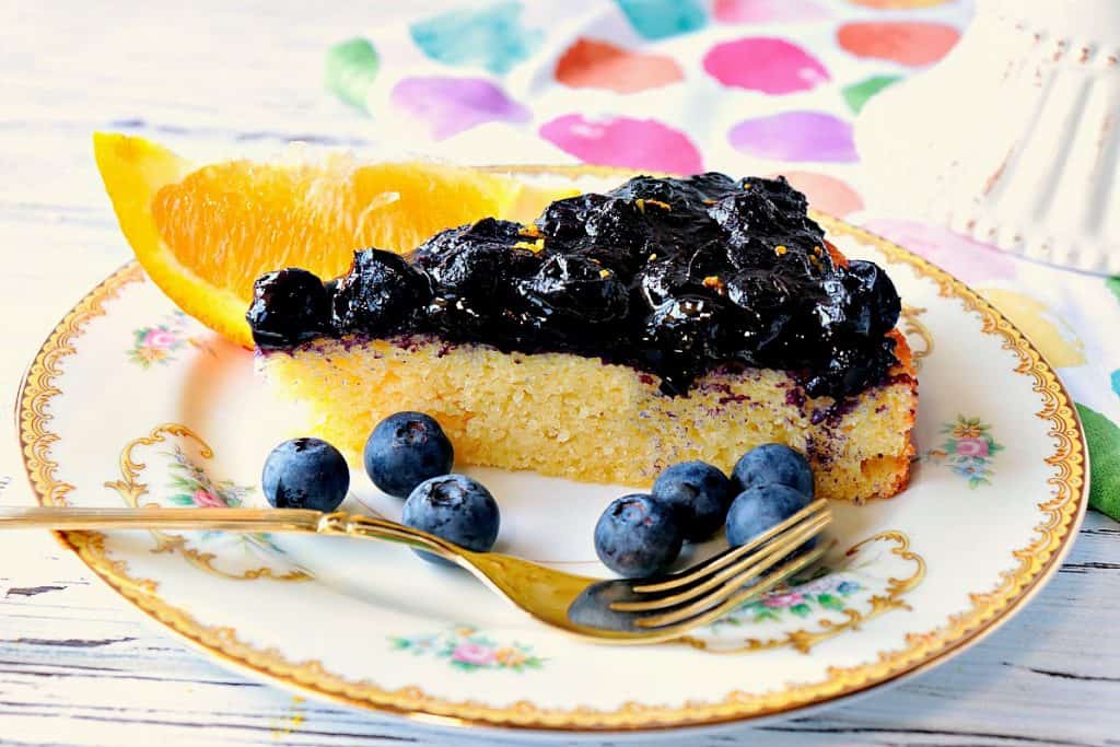 Slice of almond cake on a china plate with fresh blueberries and an orange slice.