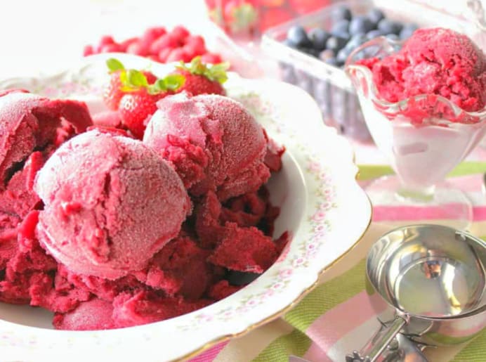 Petty bowl with scoops of strawberry sorbet on the left and an glass ice cream dish with a scoop of sorbet on the right with berries in the background.