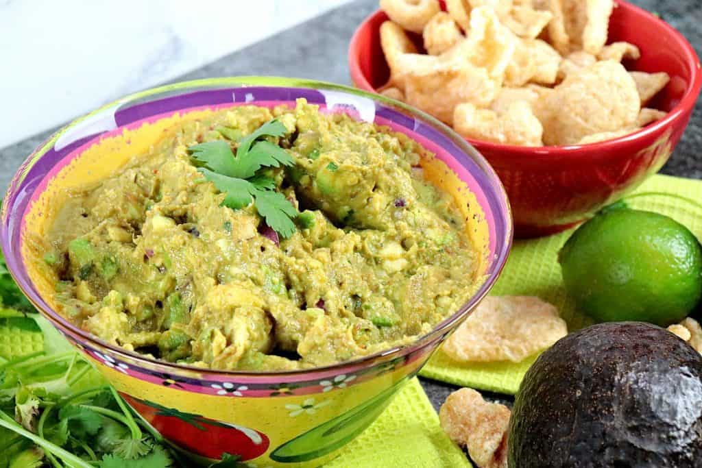 Guacamole in a colorful bowl with cilantro and pork rind dippers in the background.