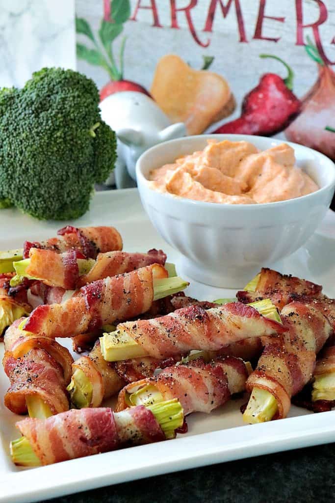 Vertical closeup image of a pile of bacon wrapped broccoli stalks on a white plate with a bowl of dipping sauce and some broccoli florets in the background.