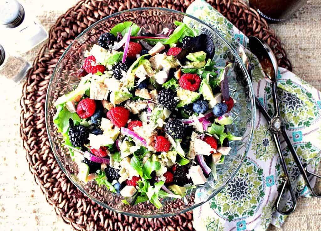 Overhead photo of a colorful berry avocado salad in a glass bowl with tongs and a salt and pepper shaker