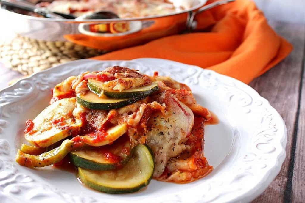 Layered zucchini parmesan on a white scalloped plate with a skillet in the background and an orange napkin.