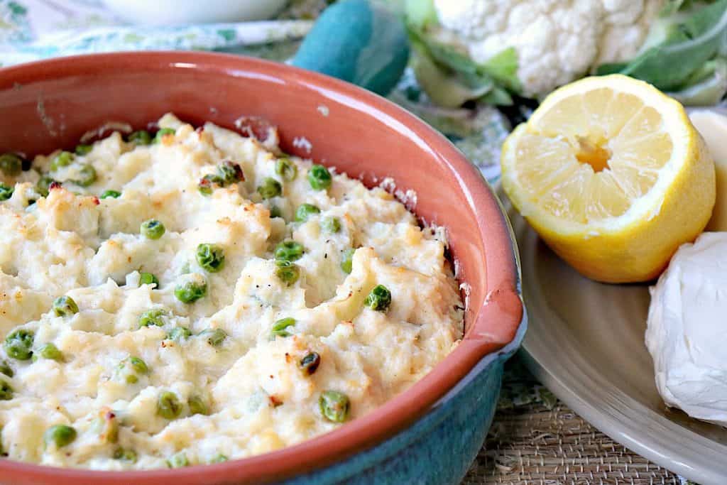 Cheesy Mashed Cauliflower Casserole in a round casserole dish with lemon and peas.