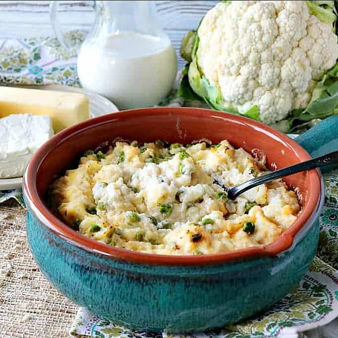 A blue casserole filled with cheesy mashed cauliflower with peas. Butter, cream, and a head of cauliflower are in the background.